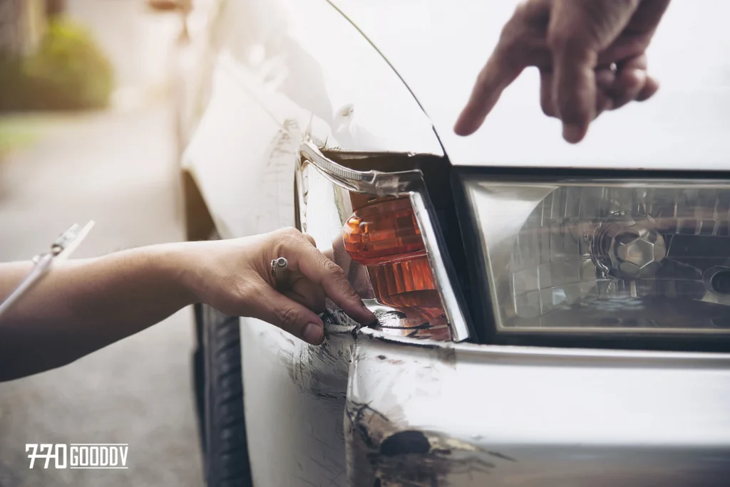 A close-up of a person's hand pointing at the dented and scratched front corner of a white car, highlighting the concept of diminished vehicle value post-accident.